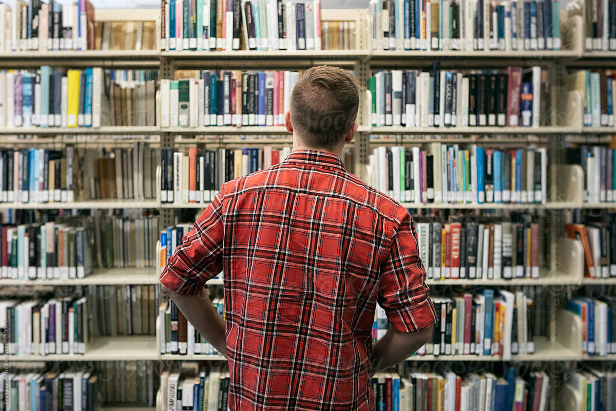 Male Student Looking At Bookshelves In Library