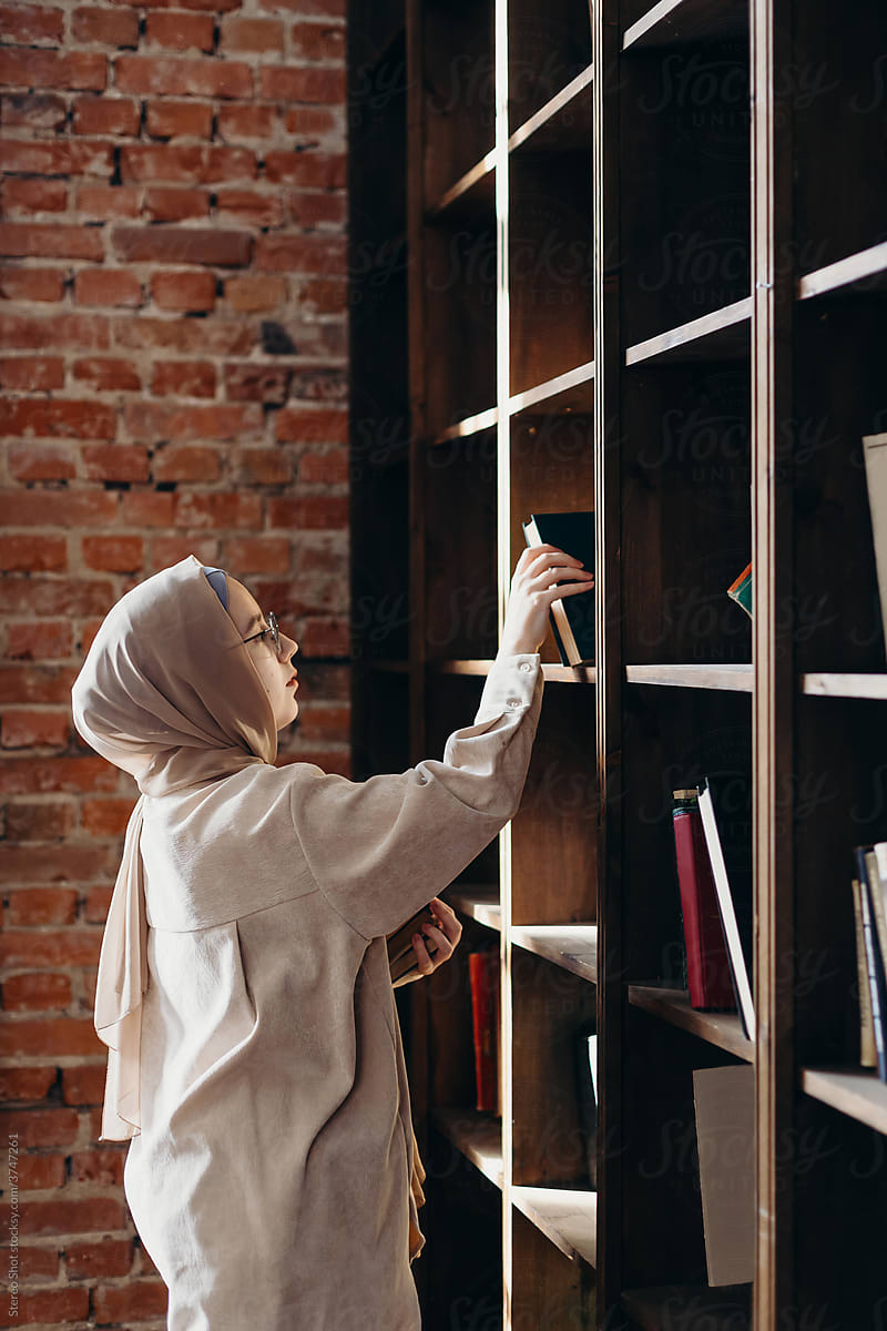 Young Muslim female student taking books from library bookshelf