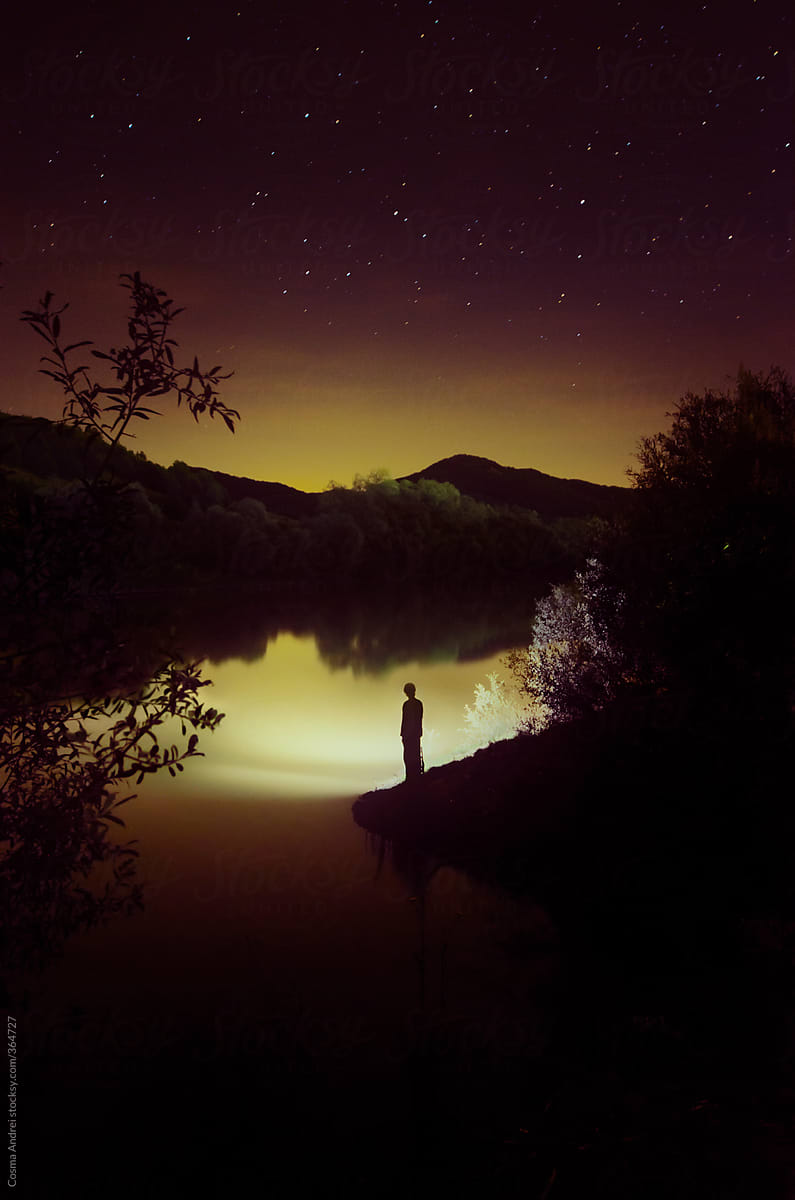 Man on lake edge at night with stars above and flashlight