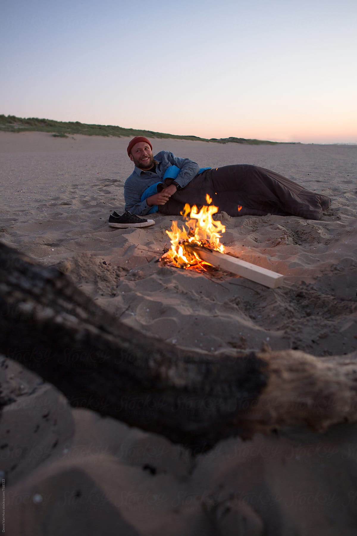 Young man laying next to bonfire on the beach in a sleeping bag.