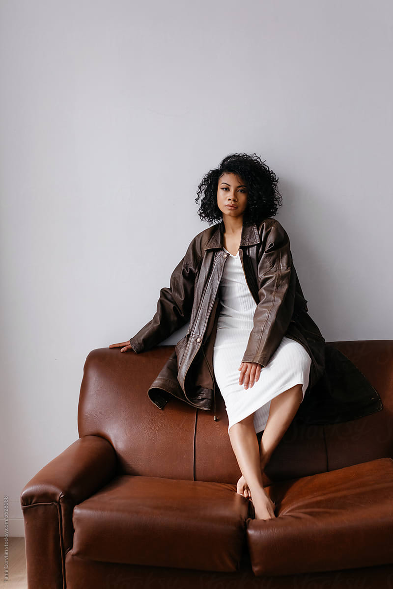 Stylish black woman in leather trench on sofa