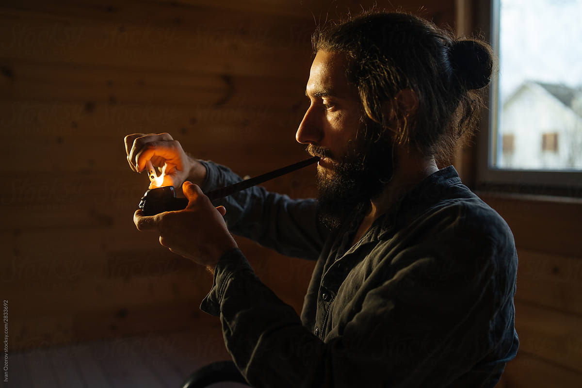 bearded man lights a pipe with a lighter in a dark room