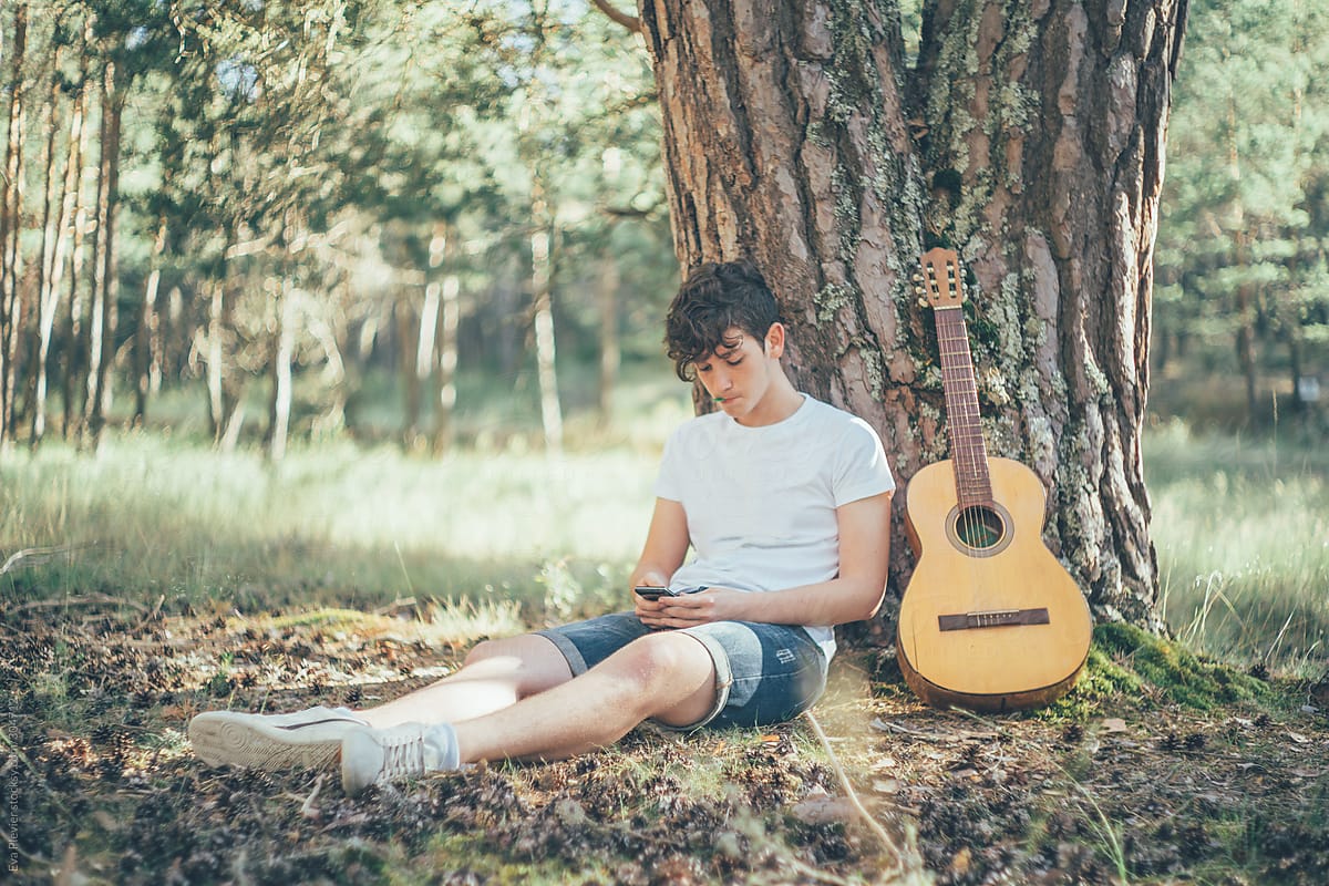 Teenage boy sitting under a tree with a guitar and texting with his cell phone.