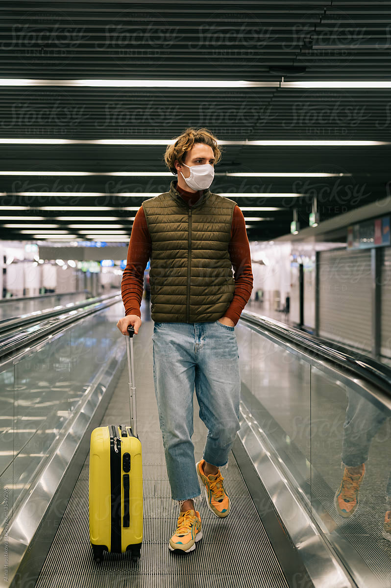 Male traveler with suitcase riding airport escalator during pandemic