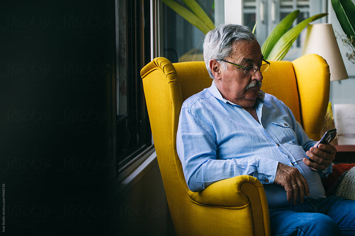 Senior male in eyeglasses using smartphone in armchair