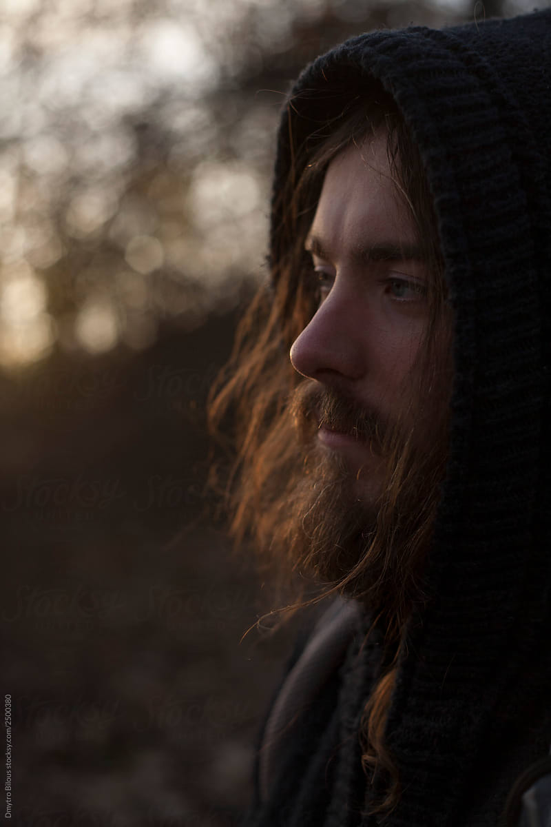 portrait of a guy in a hood with long hair and a beard at sunset