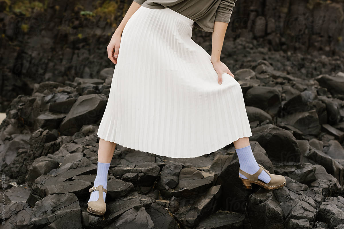 young woman in long white skirt and colored socks standing and leaning on dark jagged rocks