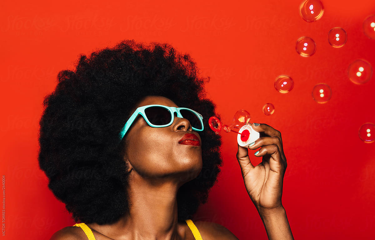 Beautiful Afro Woman on red background with blue sunglasses and blowing bubbles