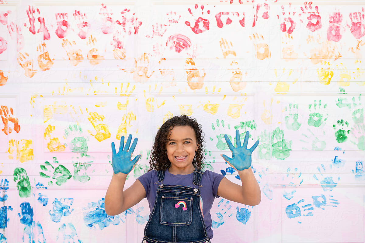 Girl with blue painted hands stands in front of rainbow mural