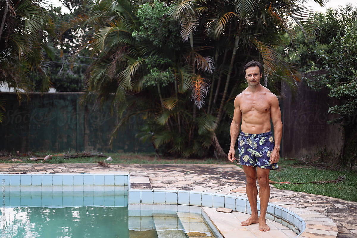 A muscled man standing next to the swimming pool and the palm trees