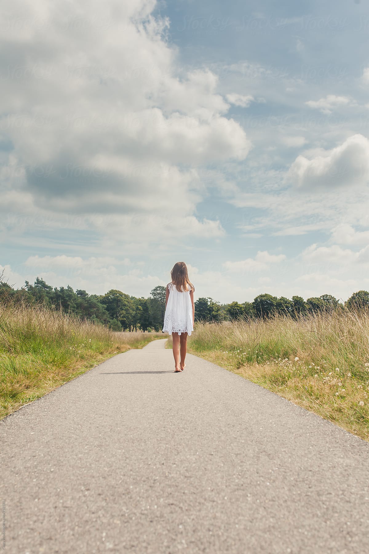 Little girl barefoot in a white dress walking on a long road