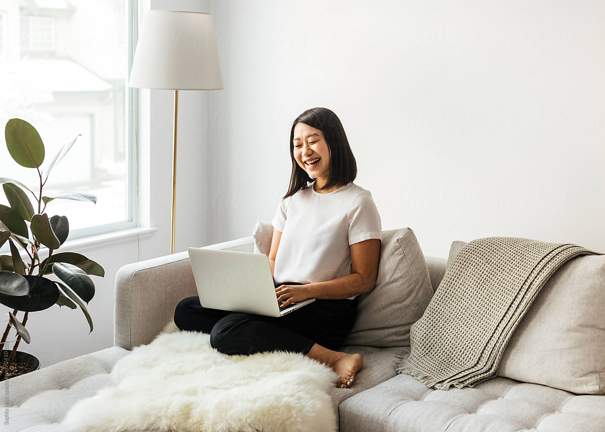 Smiling Asian female sitting on sofa working from home in minimalist space