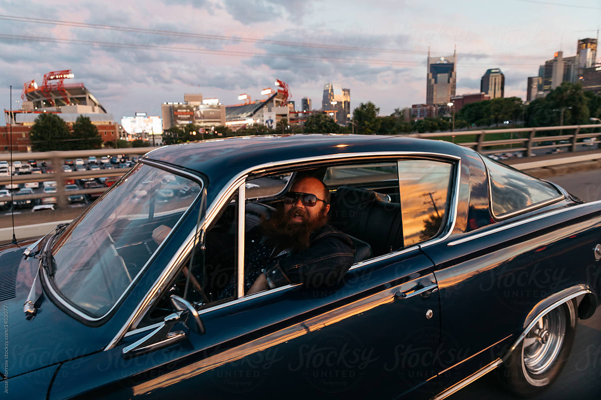 male with beard wearing sunglasses driving classic vintage retro car at sunset in city urban environment