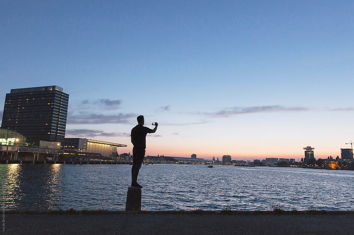 Man making a photo of the waterfront in Amsterdam, with a beautiful colourful sky. Photo taken during sunset or dusk in the summer. with a beautiful sky in the background.