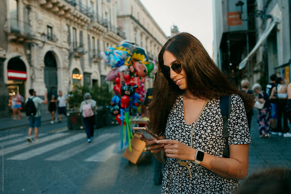 woman with sunglasses in sunlight in city street checking her phone
