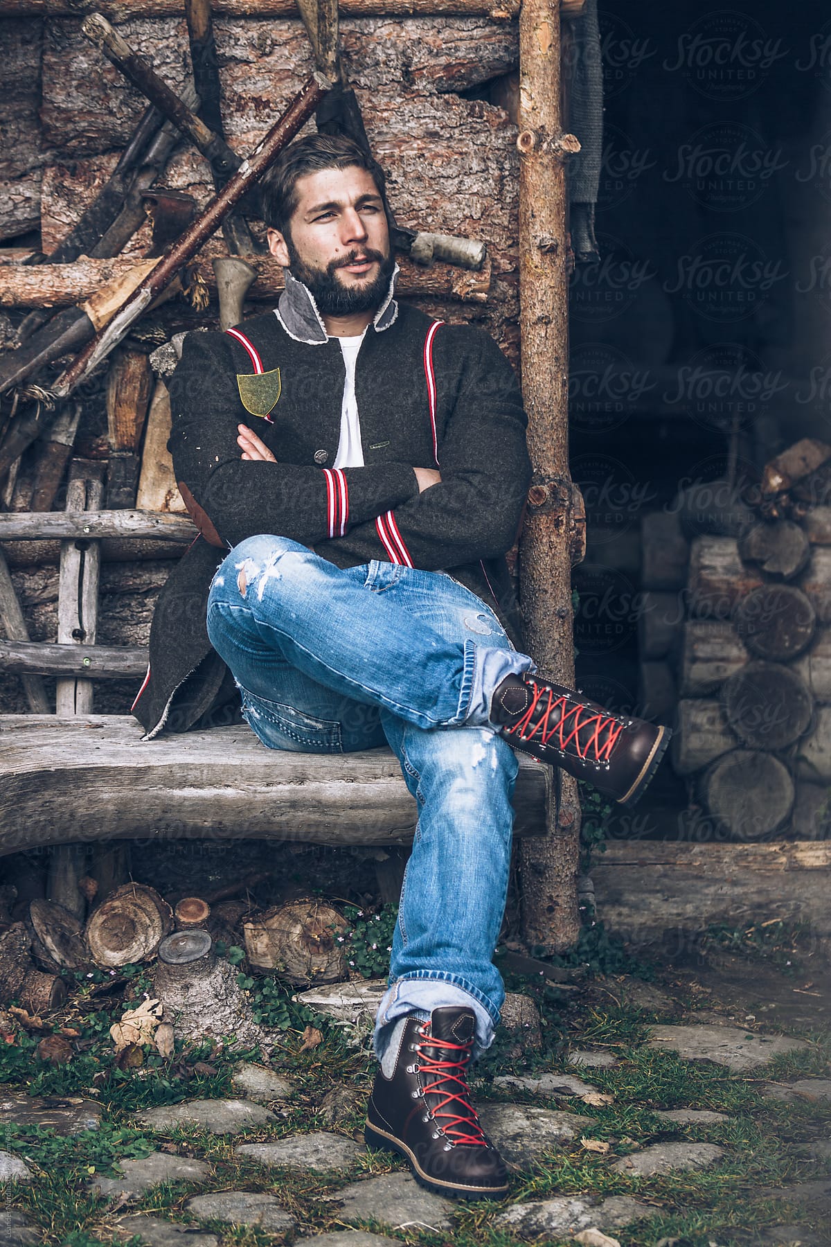 young bearded man sitting in front of an old rustic burk mulch cabin