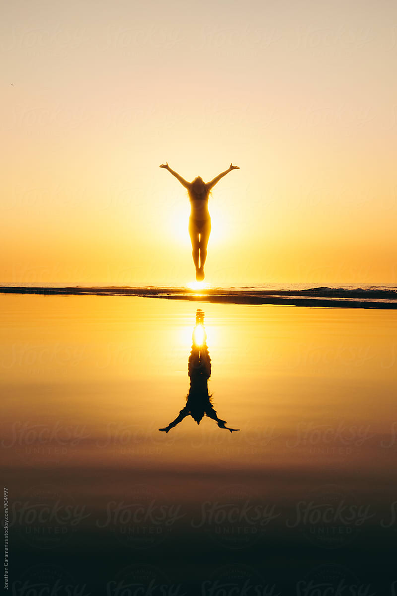 Young woman jumps for joy silhouetted by golden light at sunrise sunset with reflection in water