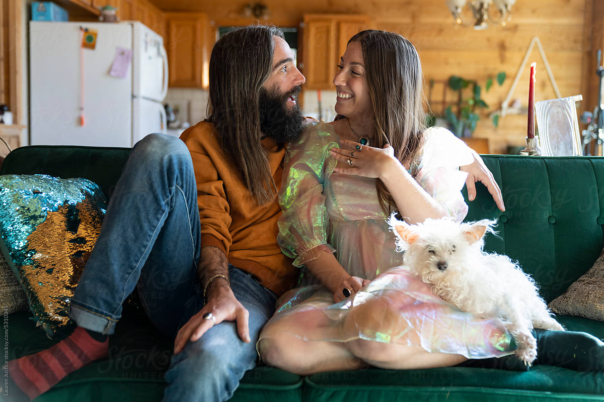 Couple in love sits with dog on green couch holding engagement ring