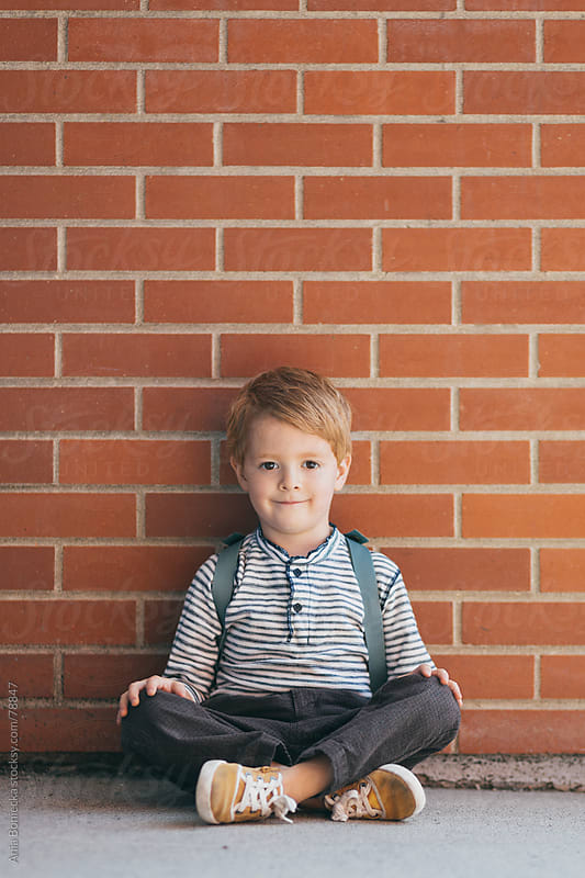 A blonde boy sitting against a brick wall with his legs crossed by Ania