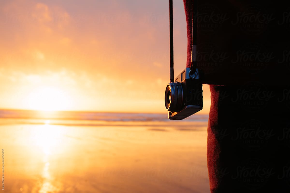 A Camera Hangs At The Side Of A Person Watching The Sun Set Over The Pacific Ocean From The Shore