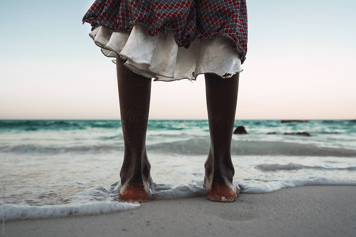 Feet of a woman standing on the sand looking towards the sea. Th
