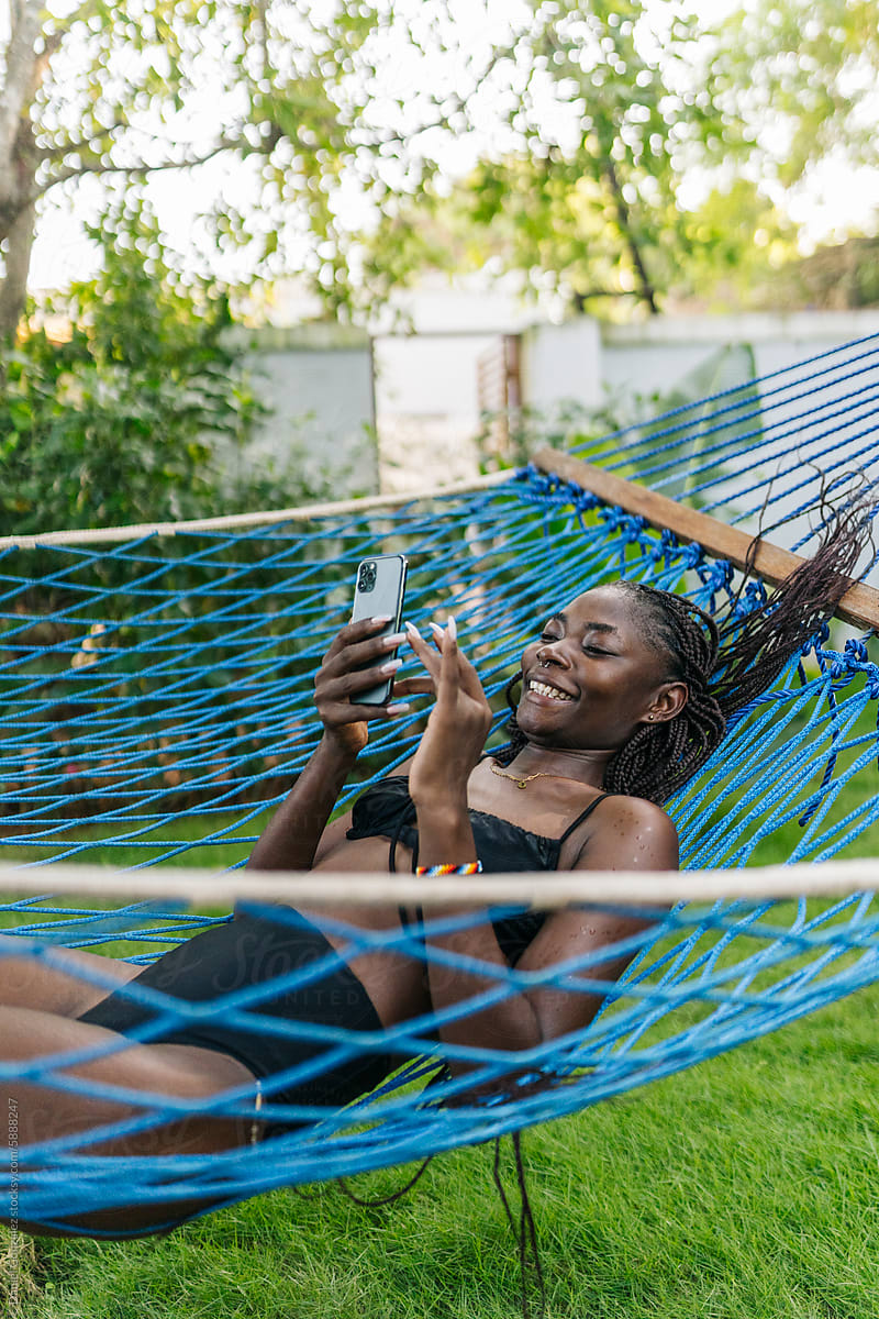 Happy young black woman surfing mobile phone while lying on hammock