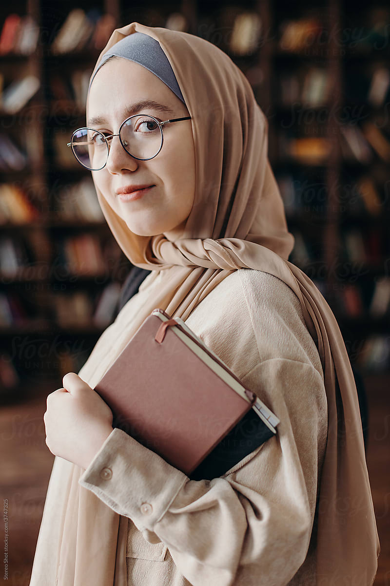 Content young Muslim female student standing with book in library