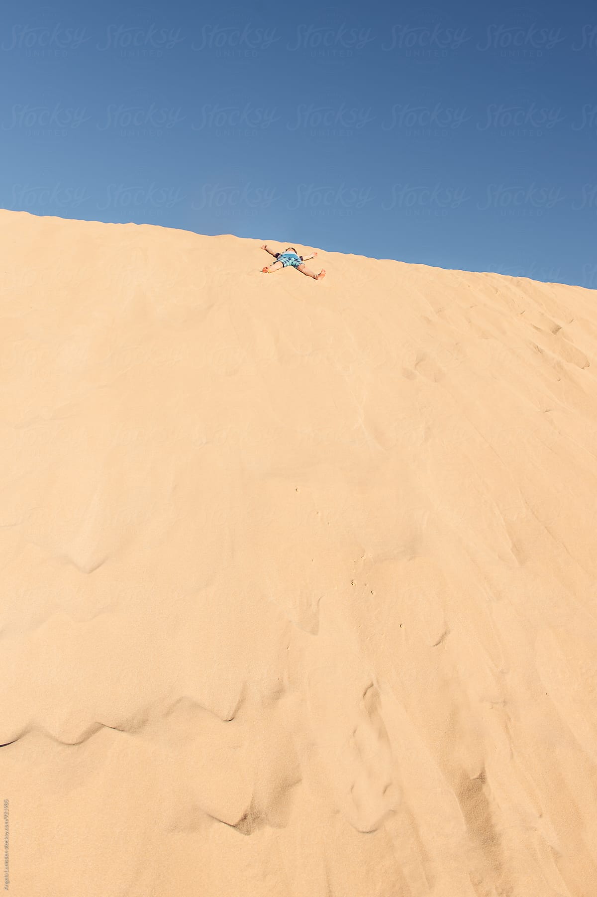 Boy doing a sand angel at the top of a large sand dune against a blue sky