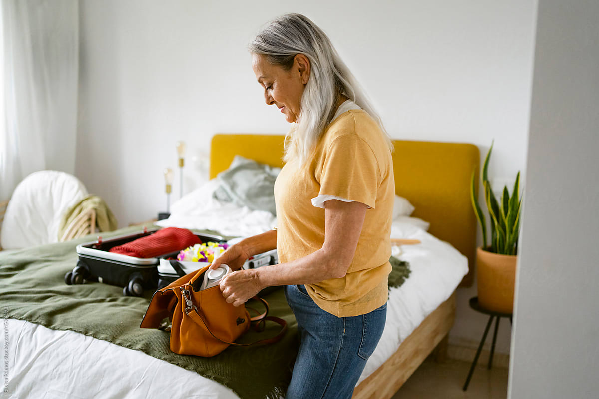 Gray-haired woman preparing travel handbag on bed