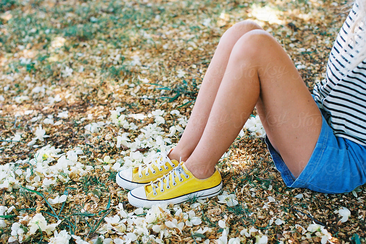 blonde woman laying in grass with flowers in striped tee and underwear and jean skirt and yellow tennis shoes