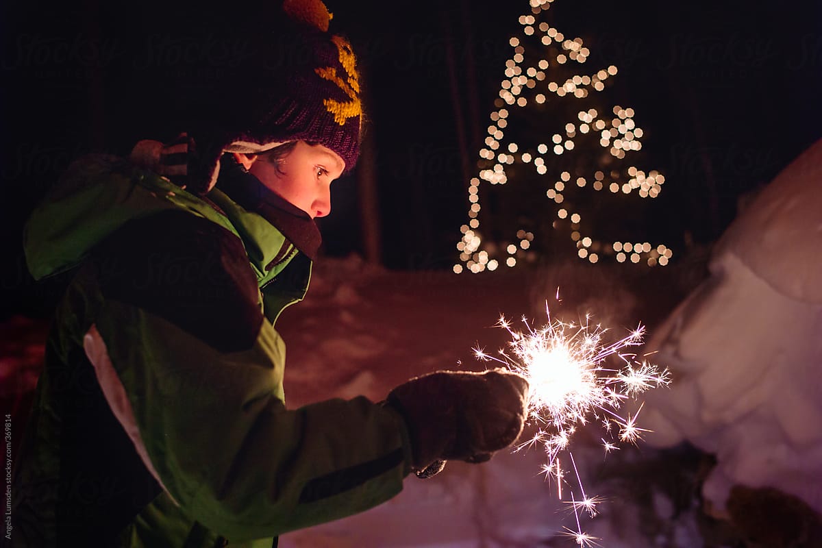 Boy in snow clothes holding a sparkler in the snow at night with holiday lights in the background