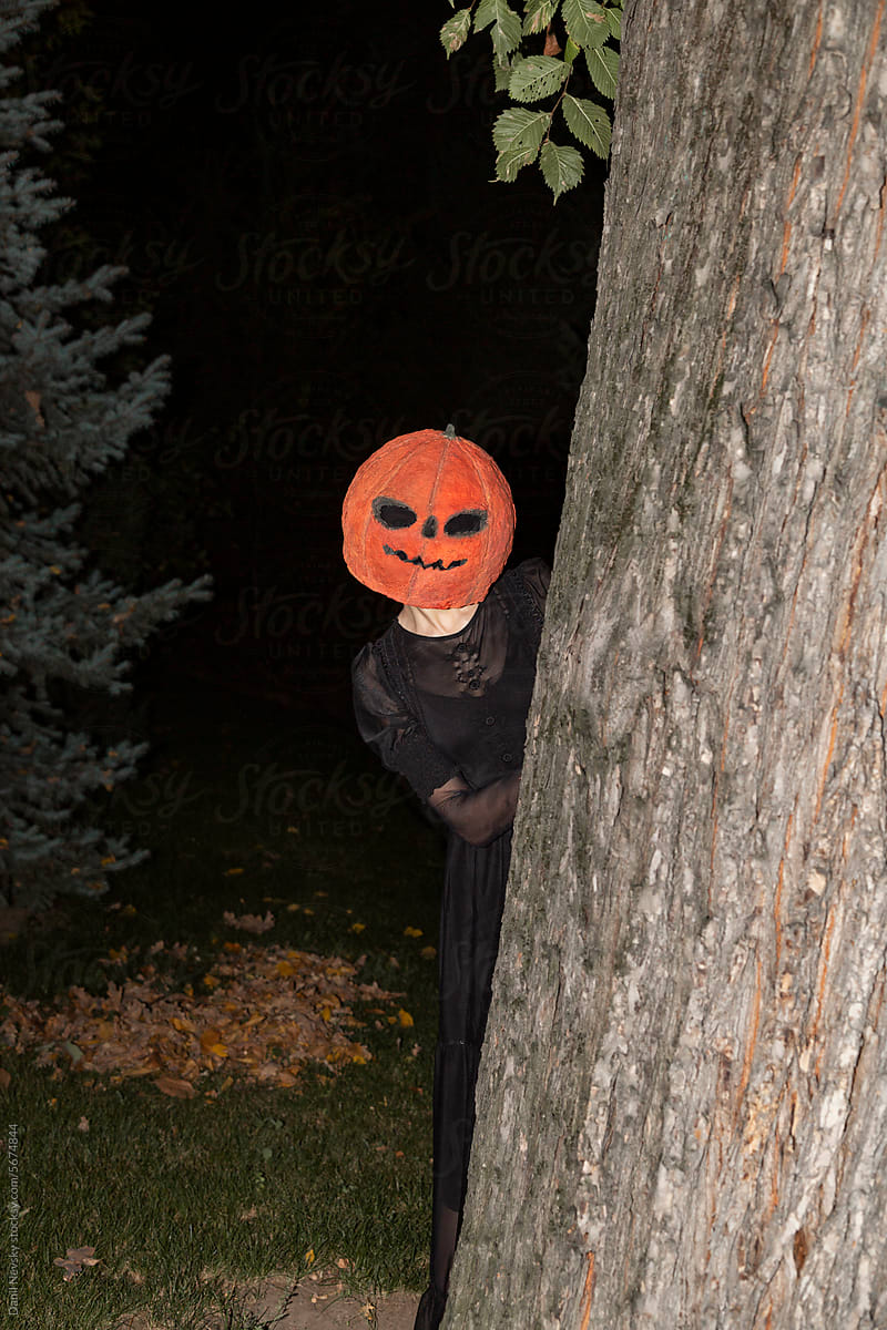 Woman with pumpkin on head peeking out from behind tree