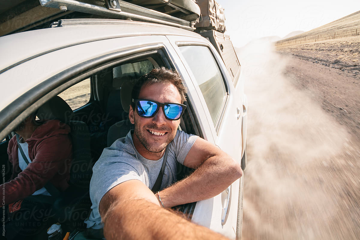 Adventure travel - Young man taking a selfie through the window of an off road car while driving on a dirt road during a road trip
