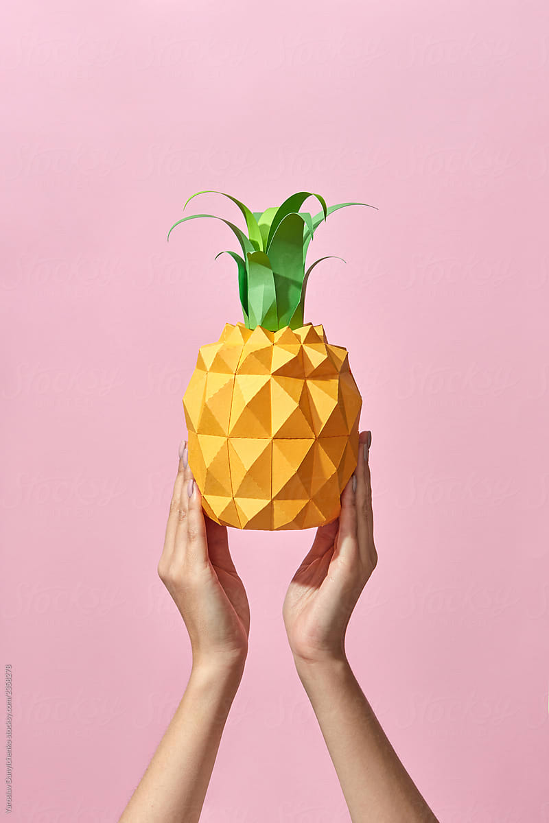 Pineapple with green leaves made of paper holds the hand of a girl on a coral background with space for text. Handcraft Tropical Fruit. Pantone 16-1546 Living Coral is the color of the year 2019.