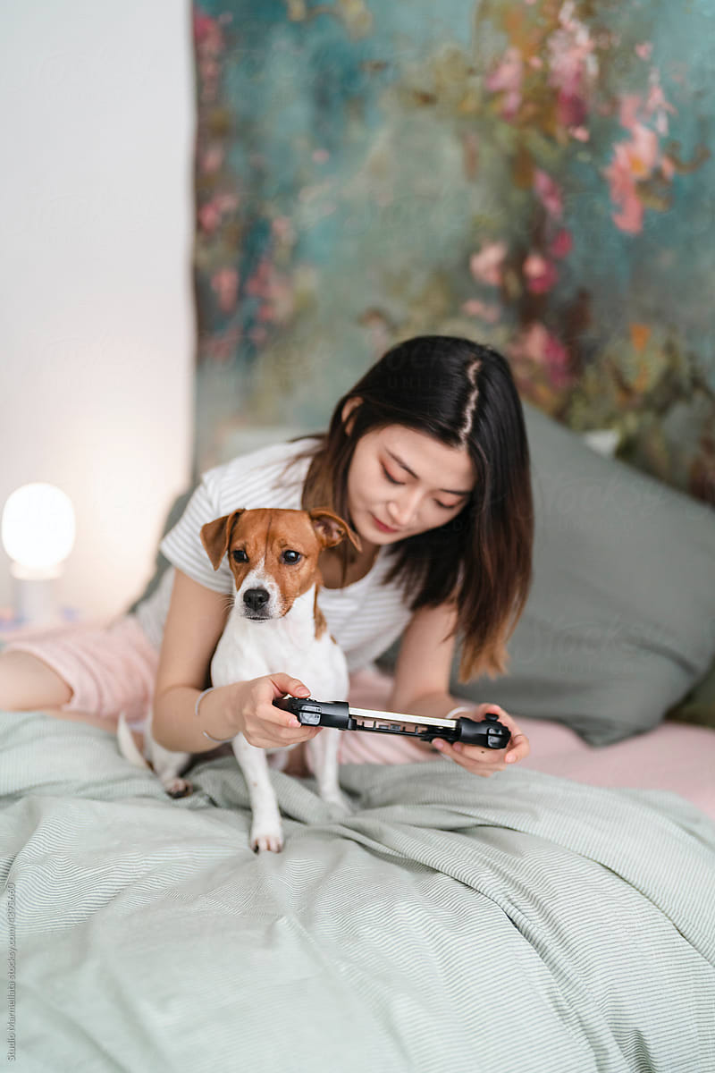 Woman playing videogame on bed with dog