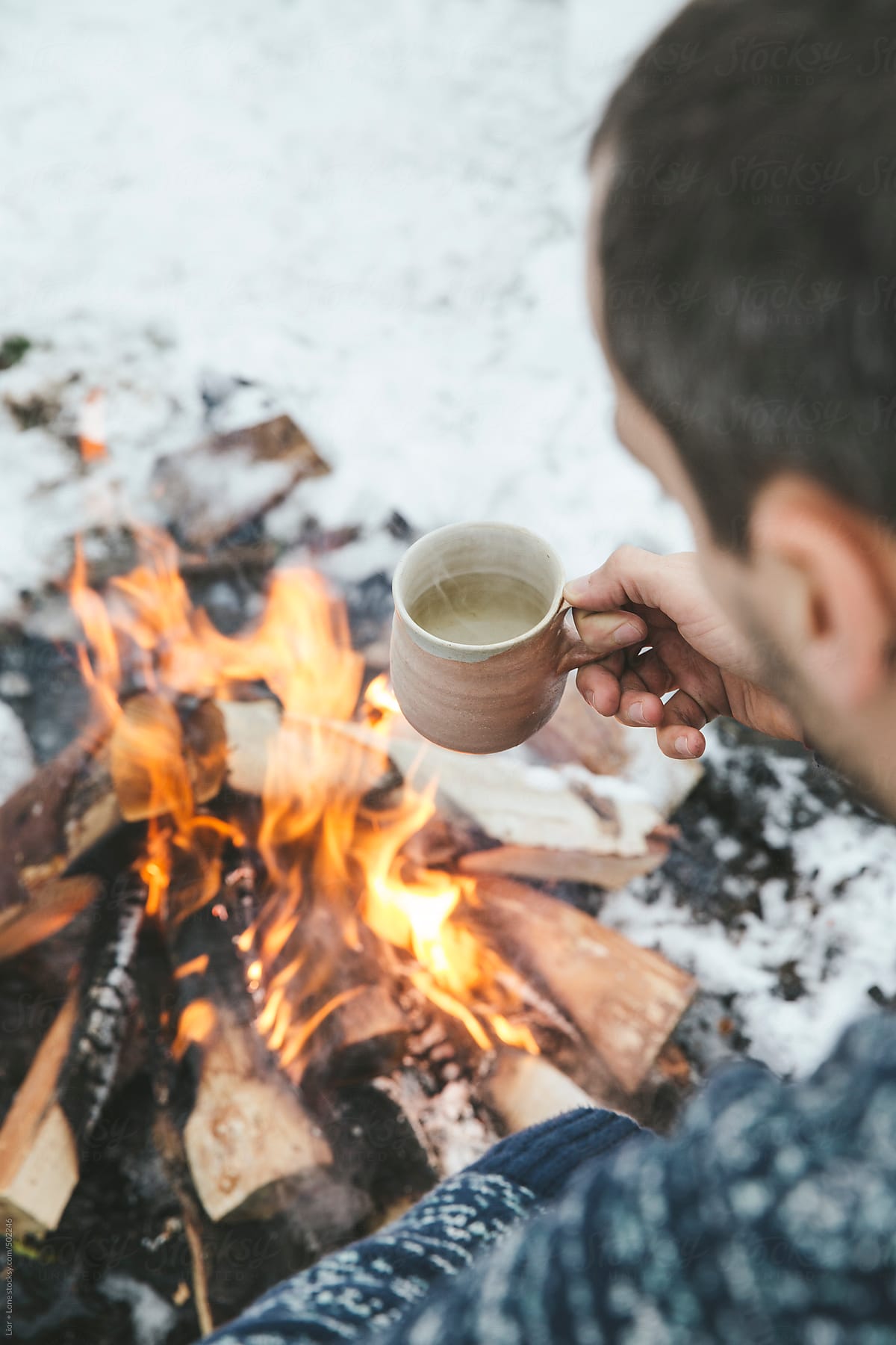 Man holding a cup of hot tea next to bonfire in the snow