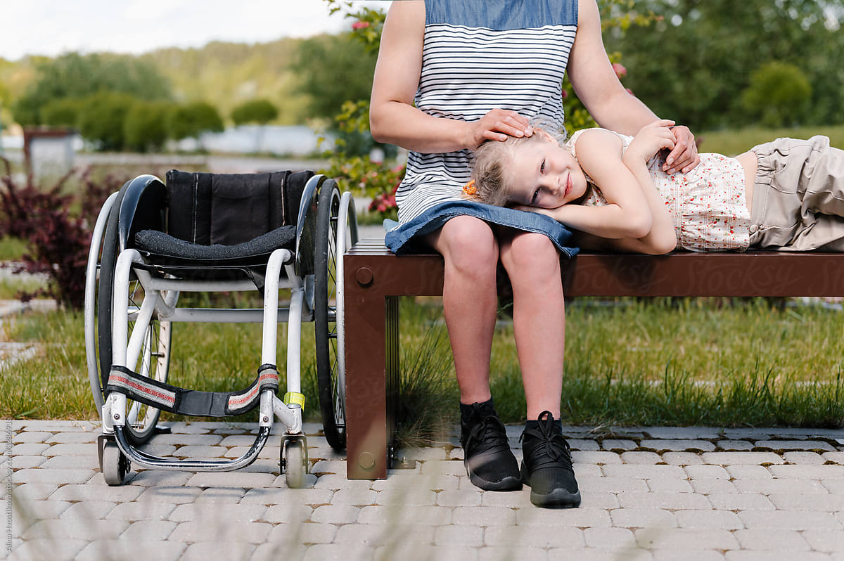 Cheerful girl lying on mothers laps on bench near wheelchair