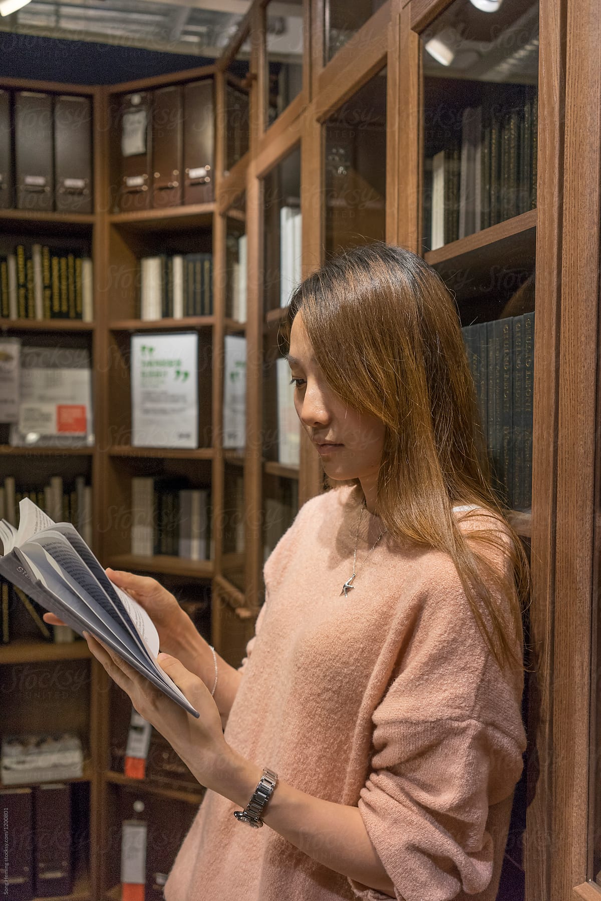 Beautiful Chinese young woman reading book against bookshelf
