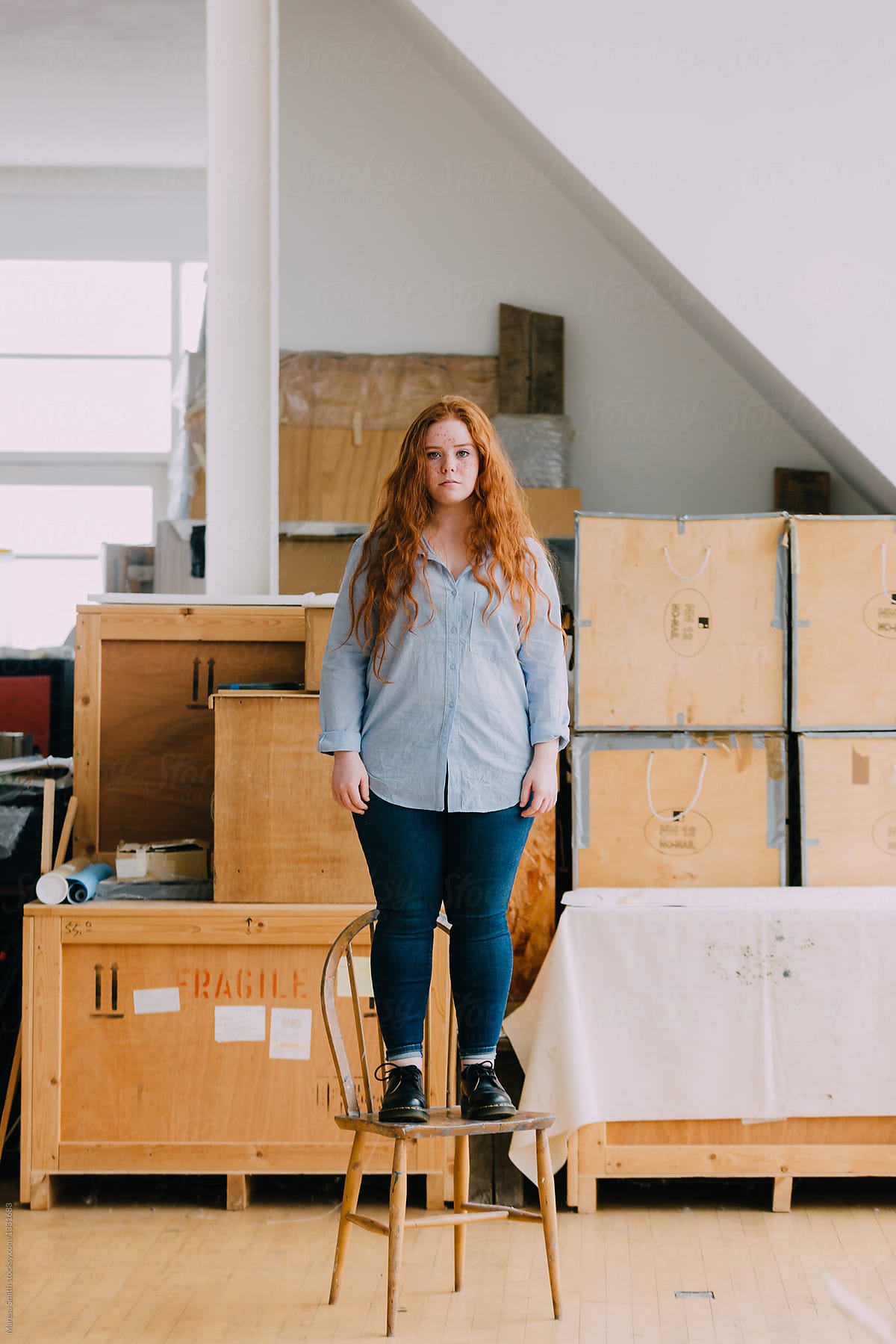 A teenage girl with ginger hair standing on a chair in a studio space surrounded by wooden boxes