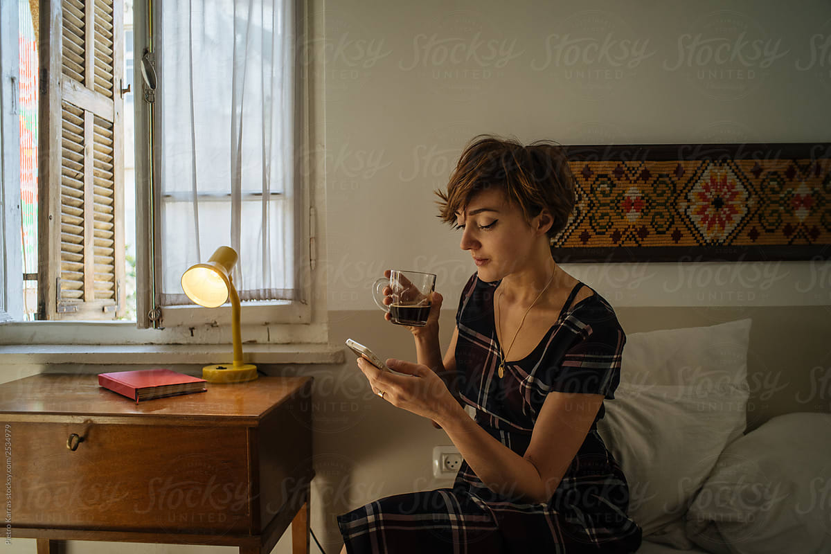 Woman with drink using smartphone on bed