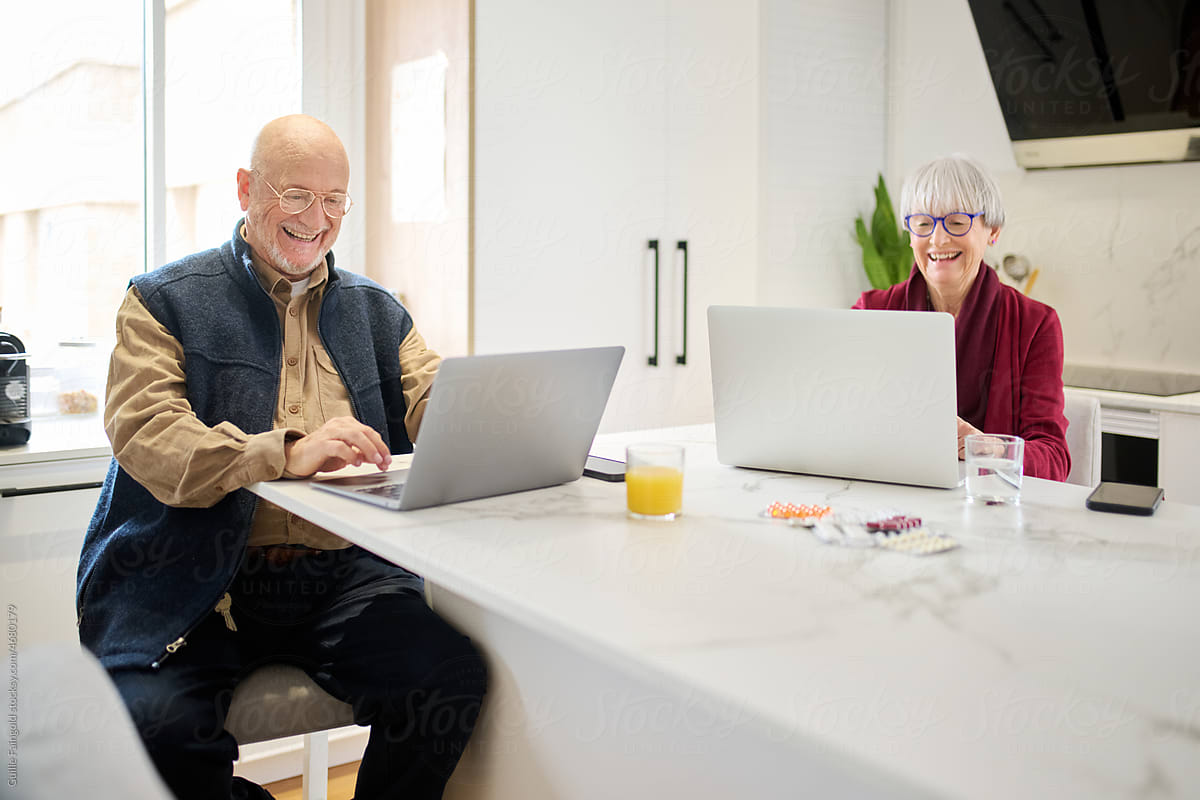 Two senior people using laptops in the kitchen.