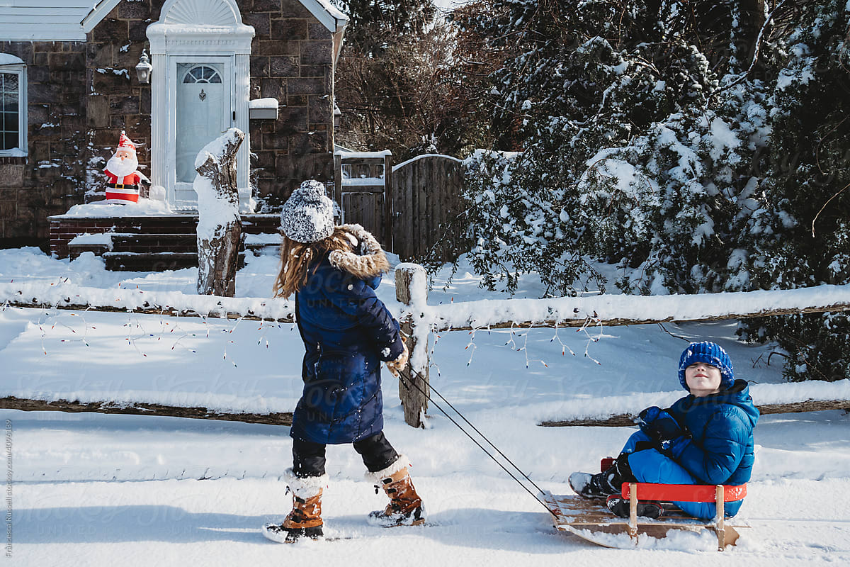 A girl pulls a boy on a sled down the snowy sidewalk.