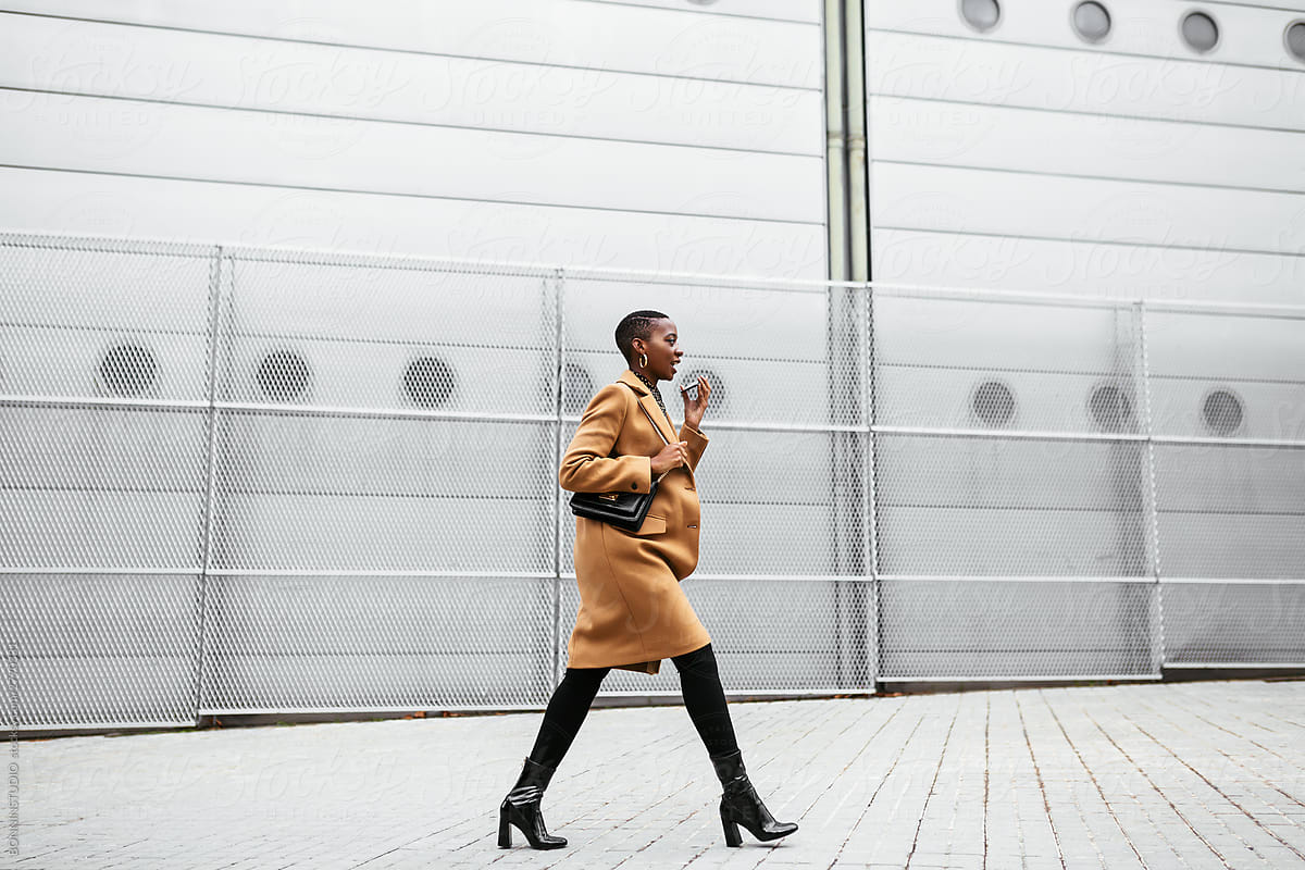 Trendy black woman speaking on smartphone and walking along urban street