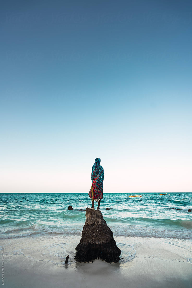 silhouette of a woman with her back on a log trunk facing the turquoise water beach