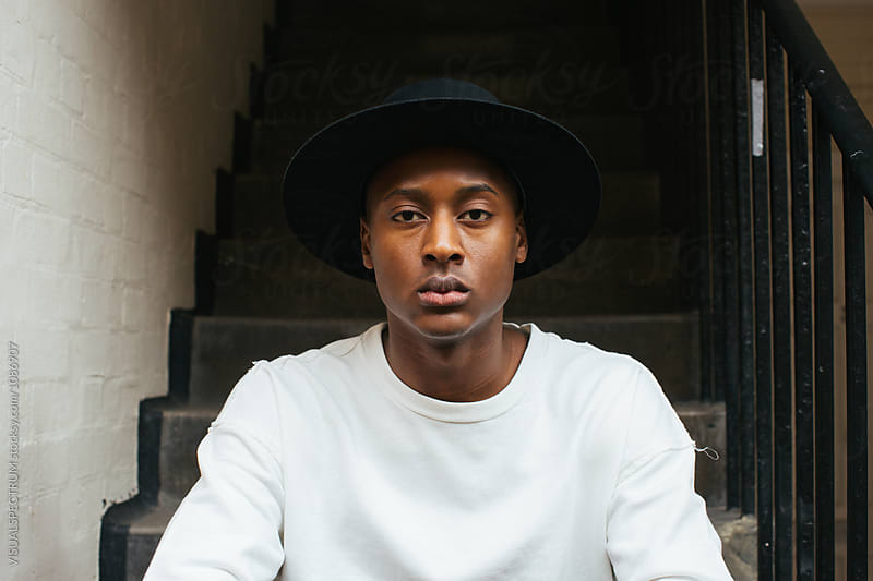 London Street Style - Outdoor Portrait of Young Cool Black Man Sitting on Stairs and Wearing Black Hat