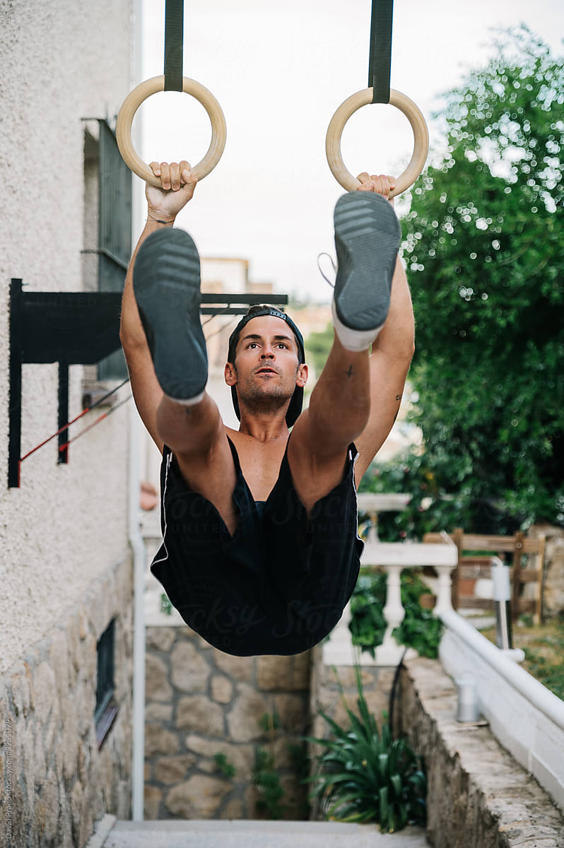 Athletic young man exercising on gymnastic rings near house