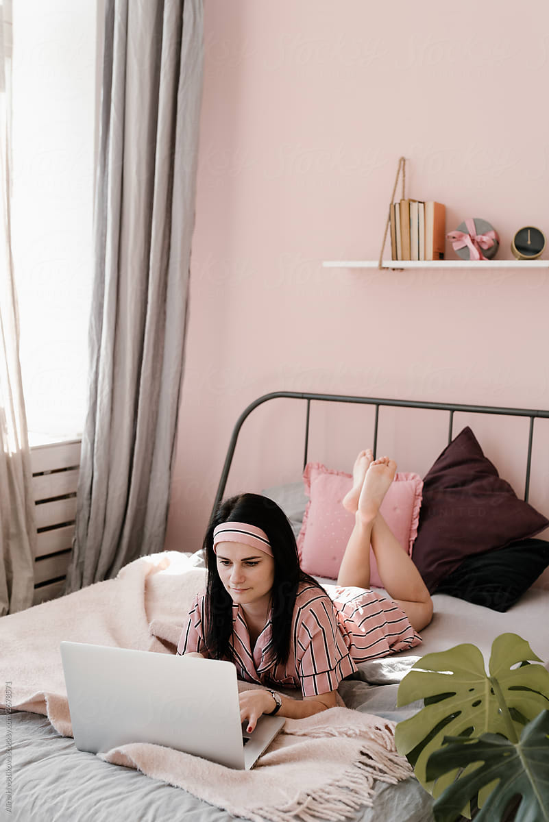 Girl wearing pink pajama lying on bed and using laptop.