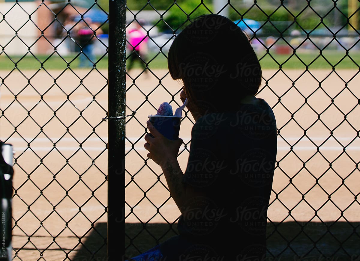 Silhouette of younger sibling eating snow cone while watching softball game