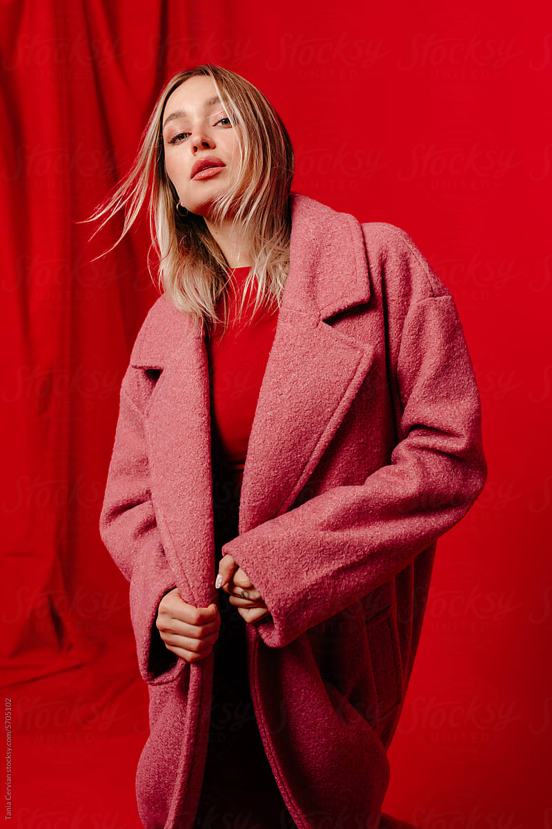 Stylish young woman standing in coat near red backdrop