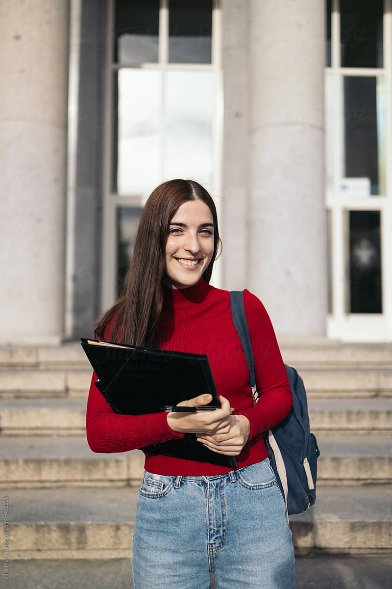 Portrait of a smiling young student with backpack and folder outdoors